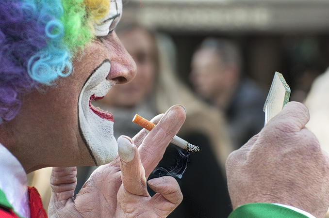 A clown looking at his makeup in a mirror while smoking in front of the Pompidou Center.