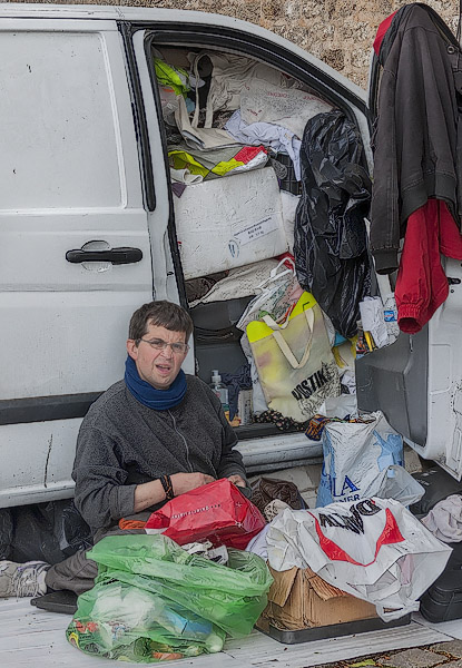 A homeless sitting in front of his van overflowing with trash, nonsense and junk.