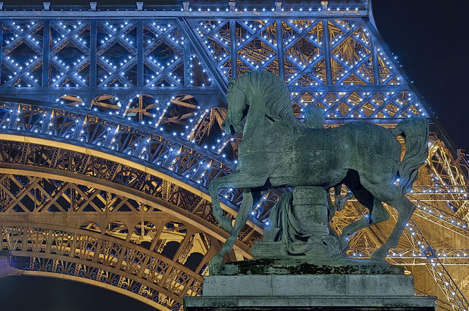 An equestrian statue of a Greek warrior in front of flashing white lights on the Eiffel Tower at night.