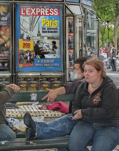 Three homeless people in front of a advertising poster for trendy spots and restaurants in Paris.